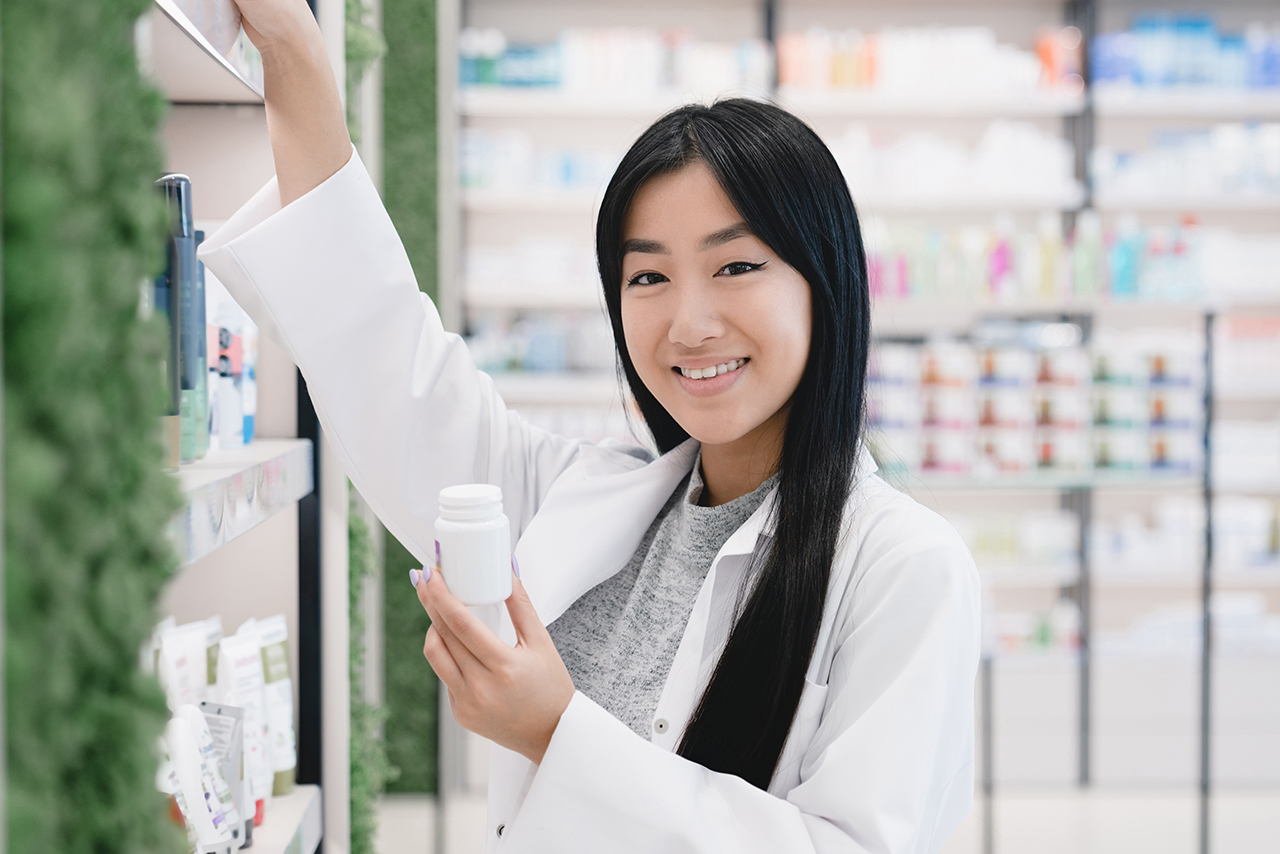 Female young asian pharmacist druggist in white medical coat holding remedy pills while looking for another medicines on shelves of pharmacy drugstore looking at camera